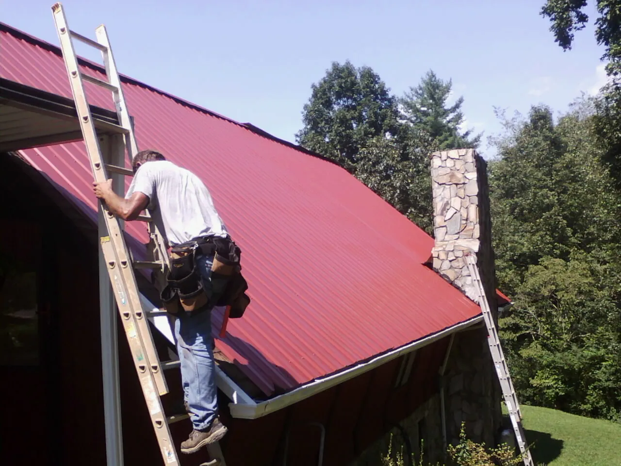 Roofer on a ladder beside a red metal roof and stone chimney — Soffit & Fascia Repair by Roofing Brothers in Hope Valley